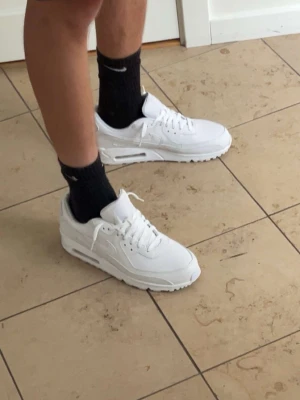 Nike Air Max 90 vita sneakers - Fräscha Nike Air Max 90 sneakers i helvitt med klassisk siluett och synlig Air-enhet i sulan. Ovandelen är i en mix av läder och mesh för både stil och komfort. Perfekta för dig som gillar en clean och sportig look. Ruggigt sköna