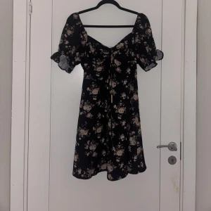 Svart blommig kortklänning med puffärm - Supersöt svart kortklänning med ljusa blommor och puffiga kortärmar. Klänningen har en v-ringad urringning och lätt volang nertill. Perfekt för dig som gillar romantisk stil och vill sticka ut lite extra.