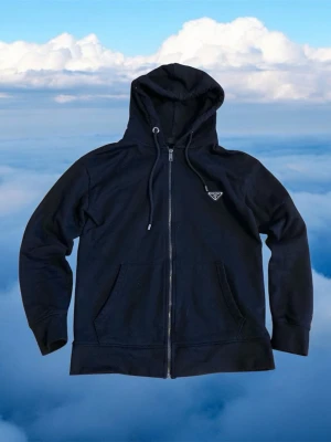 Svart hoodie med dragkedja från Prada - Snygg svart hoodie från Prada med dragkedja framtill, två fickor och justerbar huva. Klassisk triangel-logga på bröstet i metall. Tillverkad i mjukt bomullsmaterial som känns lyxigt och bekvämt. Perfekt för en stilren och avslappnad look.