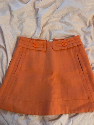 Orange kortkjol med fickor och knappar - Snygg orange kortkjol i A-linjeform med dekorativa knappar framtill och praktiska fickor både fram och bak. Kjolen har en dragkedja bak och är tillverkad i ett strukturerat tyg som ger en cool retrovibe. Perfekt för dig som vill sticka ut med färg! Kjolen är i stl 40 men skulle mer säga att den är i stl 34-36 (xs/s) så jag själv har s men är lite liten i midjan
