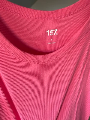 Rosa ribbad ärmlös klänning 157 - Snygg ärmlös klänning från 157 i en stark rosa färg. Klänningen är ribbad och har en rund halsringning. Tillverkad i 100% bomull vilket gör den både bekväm och luftig. Perfekt för varma dagar och enkel att styla med sneakers eller sandaler. 