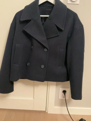 Mörkblå skepparkavaj med bred krage - Stilren mörkblå skepparkavaj med dubbelknäppning och breda slag. Jackan har raka fickor framtill och en boxig, något kortare passform. Perfekt för dig som gillar klassisk och tidlös stil. Materialet ser ut att vara ullblandning.