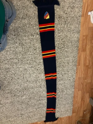 Djurgården halsduk i blått och rött - Snygg halsduk i mörkblått med röda och gula ränder samt DIF-logga broderad på ena sidan. Halsduken har fransar i ändarna och är perfekt för att visa ditt stöd på matchen. Materialet är stickat och mjukt.