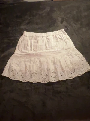 Vit kortkjol - Söt vit kortkjol med resår i midjan och volangkant. Kjolen har broderade blommönster längs nederkanten. Perfekt för varma dagar och enkel att matcha med olika toppar.