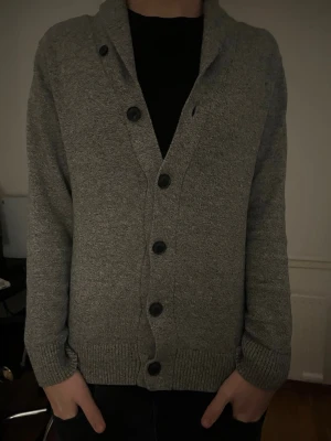 Grå kofta  - Snygg grå kofta med stora svarta knappar och ribbade muddar. Koftan har en klassisk krage och är tillverkad i ett mjukt, stickat material. Perfekt att slänga på sig över en t-shirt för en avslappnad look. Väldigt skönt material! Modellen är 180 
