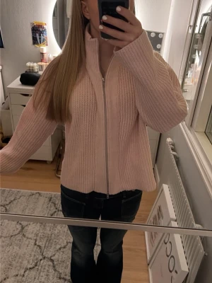 Ljusrosa ribbstickad ziptröja - Mysig ljusrosa ribbstickad tröja med dragkedja framtill och hög krage. Tröjan har en avslappnad passform och långa ärmar, perfekt för lager-på-lager-stil. Enkel och stilren design som passar till många outfits.