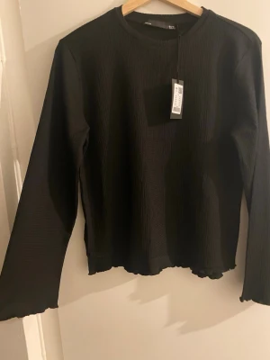 Svart ribbad långärmad topp 157 - Svart långärmad topp från 157 med ribbad struktur och rund hals. Toppen har en enkel och clean design med lätt vågiga kanter vid ärmslut och nederkant. Perfekt att styla med jeans eller kjol för en trendig look ✨