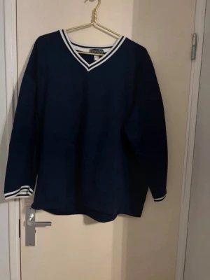 Mörkblå stickad tröja med V-ringning - Mörkblå stickad tröja från Ravens med vit rand vid V-ringad hals och ärmslut. Tröjan har en klassisk och sportig look med långa ärmar och ribbade detaljer. Perfekt för lager-på-lager och enkel att matcha med jeans eller kjol.