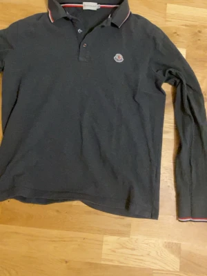 Mörkgrå Moncler pikétröja - Snygg mörkgrå långärmad pikétröja från Moncler med klassisk krage och knappar framtill. Tröjan har Moncler-logga på bröstet och diskreta röda och vita detaljer på krage och ärmslut. Perfekt för en stilren och avslappnad look.