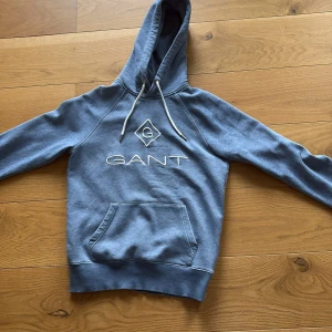 Blå hoodie från Gant - Säljer en blå hoodie från GANT i storlek XS. Tröjan har vit broderad logga framtill, huva med snörning och en stor magficka. Tröjan har ett pyttelitet hål på framsidan men som bara syns på nära håll. 