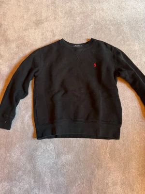 Svart seatshirt från Polo Ralph Lauren - Klassisk svart sweatshirt från Polo Ralph Lauren med rund halsringning och röd broderad logga på bröstet. Tröjan har långa ärmar och ribbade muddar vid ärmslut och nederkant. Perfekt för en stilren och avslappnad look. Ny pris 1200kr