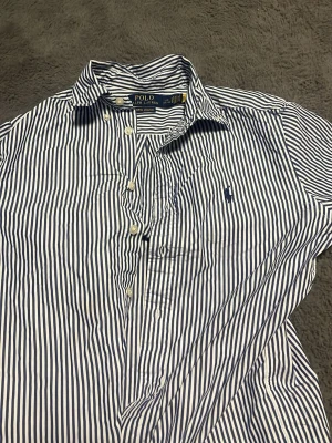 Randig skjorta från Polo Ralph Lauren - Snygg skjorta från Polo Ralph Lauren i vitt och blått med klassiskt randigt mönster. Skjortan har lång ärm, knappar framtill och broderad logga på bröstet. Tillverkad i bomull med lite stretch för extra komfort.