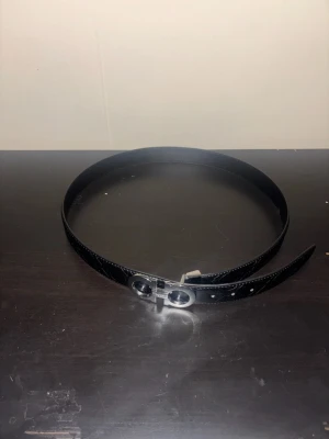 Svart skärp från Salvatore Ferragamo - Snyggt svart skärp från Salvatore Ferragamo med ikoniskt dubbel-Gancini-spänne i silverfärg. Skärpet är gjort i slätt skinn och har präglade detaljer längs hela bandet. Perfekt accessoar för att lyfta din outfit.
