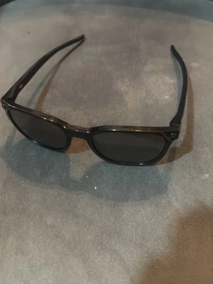 Svarta solglasögon från Oakley - Snygga svarta solglasögon från Oakley med klassisk rektangulär form och diskret logga på skalmen. Bågarna är tillverkade i lätt syntetmaterial och glasen är mörka, perfekta för soliga dagar.