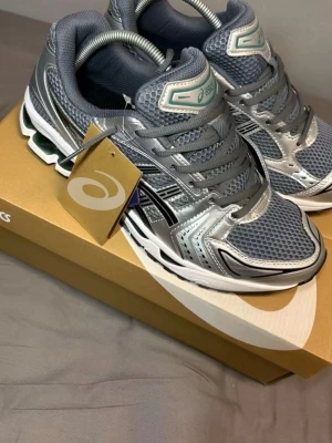 Gråa ASICS sneakers med silvriga detaljer - Snygga sneakers från ASICS i grått med silvriga och svarta detaljer. Ovandelen är i mesh och syntetmaterial för bra andningsförmåga och komfort. Sulan är vit med svarta och gröna inslag, och skorna har klassisk snörning. Perfekta för en sportig och trendig look.