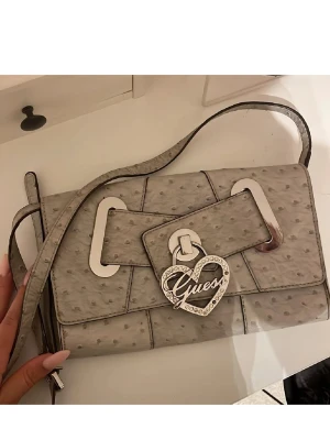 Grå axelväska från Guess med hjärtdetalj - Snygg grå axelväska från Guess i präglat material med strutsliknande mönster. Väskan har en lång axelrem, silverfärgade metalldetaljer och ett stort hjärthänge med Guess-logga framtill. Perfekt för att lyfta din outfit med en trendig touch.