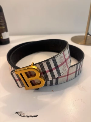 Burberry rutigt bälte med guldspänne - Snyggt bälte från Burberry med klassiskt rutigt mönster i vitt, svart och rött. Bältet har ett stort guldigt B-spänne som ger en lyxig känsla. Perfekt accessoar för att lyfta din outfit.
