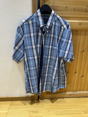 Blå rutig kortärmad skjorta - Snygg blå rutig skjorta med korta ärmar och klassisk krage. Skjortan har knappar framtill och en bröstficka. Perfekt för en avslappnad stil och tillverkad i mjuk bomull.