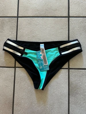 Turkos och svart bikiniunderdel Censay - Snygg bikiniunderdel från Censay i turkos och svart med vita detaljer och dekorativa svarta dragkedjor på sidorna. Modellen har en sportig look och är tillverkad i ett stretchigt syntetmaterial som passar perfekt för strand och pool.