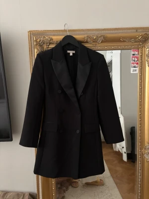 Svart kavaj klänning med satinkrage H&M - Stilren svart kavaj klänning från H&M i storlek 34 med dubbelknäppning och breda slag i satin. Kavajen har två fickor framtill och en klassisk, rak passform som ger en snygg siluett. Perfekt för dig som vill ha en tidlös och trendig look.