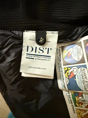 Svart dunjacka från Moncler - Cool svart dunjacka från Moncler med klassisk inneretikett och serietidningsmönster på fodret. Jackan har DIST-certifiering för spårbar dunfyllning och exklusiv känsla. Perfekt för kalla vinterdagar.