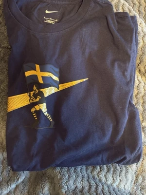 AIK vi är överallt edition - Snygg mörkblå t-shirt från Nike med ett stort guldigt Nike-logga och svensk flagga på bröstet. Tillverkad i mjuk bomull och har en klassisk passform med korta ärmar. Perfekt för dig som vill ha en sportig och stilren look. Använd 1 gång