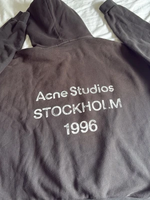 Svart hoodie Acne Studios - Snygg mörkgrå hoodie från Acne Studios med tryck på ryggen: 'Acne Studios STOCKHOLM 1996'. Klassisk passform med huva och mjukt material, perfekt för en chill och trendig look.