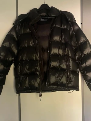 Svart pufferjacka med huva - Stilren svart pufferjacka med glansig finish och huva. Jackan har dragkedja framtill, två sidofickor med dragkedja och breda kanaler för extra värme. Perfekt för kalla vinterdagar.