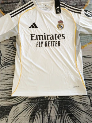 Real Madrid matchtröja Adidas vit - Säljer en vit Real Madrid matchtröja från Adidas med svarta detaljer och gula linjer längs sidorna. Tröjan har klubbmärke på bröstet, Adidas-logga och sponsortryck 'Emirates Fly Better' framtill. Tillverkad i lätt och ventilerande material med Aeroready-teknologi.