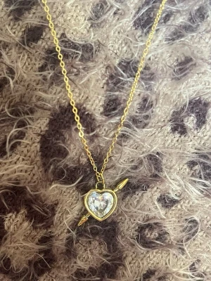 Halsband med hjärthänge och pil vit guld - Guldfärgat halsband med ett hjärthänge i metall, dekorerat med en gnistrande klar sten i mitten. Hänget har en pil som går igenom hjärtat vilket ger en romantisk och trendig look. Perfekt accessoar för att lyfta din outfit. Rostfri