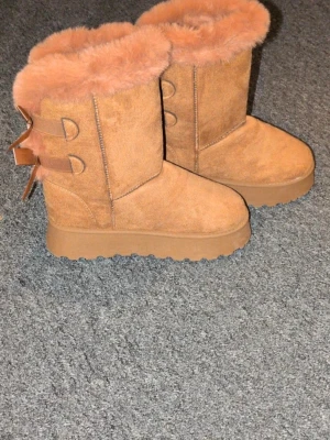 Bruna fluffiga stövlar med rosetter - Säljer ett par bruna boots med fluffigt foder och två söta rosetter bak. Skorna har en högre sula och är gjorda i mockaimitation. Perfekta för kalla dagar när du vill vara både varm och snygg. 