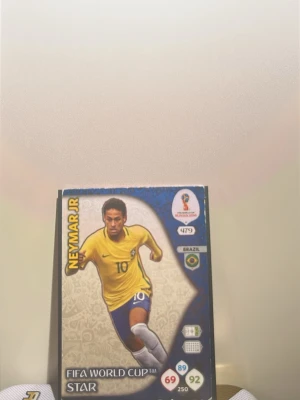 FIFA World Cup Star samlarkort Neymar Jr - Samlarkort med Neymar Jr från FIFA World Cup. Kortet har blå och gula färger, visar spelaren i Brasiliens landslagströja och har statistik samt turneringslogga. Perfekt för dig som gillar fotboll och samlar på sportkort.
