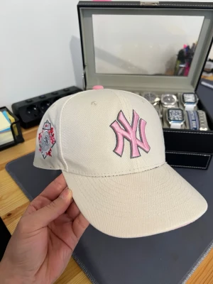 Vit New Era NY keps med rosa detaljer - Snygg vit keps från New Era med klassisk NY-logga i rosa framtill och broderad detalj på sidan. Skärmen är rosa undertill och modellen är 59FIFTY. Perfekt accessoar för en sportig och trendig look.