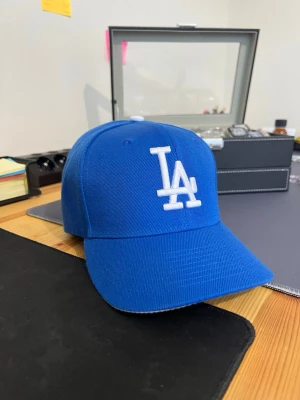 Blå LA-keps från New Era - Snygg blå keps från New Era med LA-logga broderad i vitt framtill. Klassisk böjd skärm och justerbar passform. Perfekt för dig som gillar sportig streetstyle och vill ha en ikonisk accessoar till din outfit.