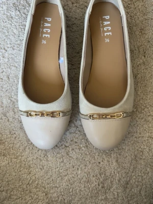 Beige ballerinaskor från Pace - Stilrena beige ballerinaskor från Pace med rund tå och guldfärgad kedjedetalj framtill. Skorna är i mocka och läder, Perfekta för dig som gillar klassisk och enkel stil. (Pris kan diskuteras)
