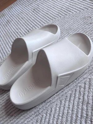 Vita tofflor från Gymshark - Säljer ett par tofflor från Gymshark. Endast använda inomhus ett fåtal gånger så är i nyskick.