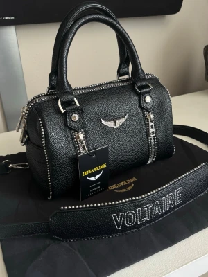 Svart handväska Zadig & Voltaire - Snygg svart handväska från Zadig & Voltaire med silverfärgade detaljer och dragkedjor. Den har en justerbar och avtagbar axelrem. Väskan är nästintill oanvänd. 