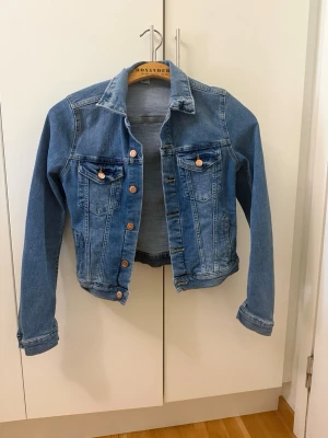 Klassisk blå jeansjacka - Snygg blå jeansjacka med klassisk passform och kopparfärgade knappar. Jackan har två bröstfickor med lock och knappar samt två sidofickor. Perfekt till lager-på-lager och enkel att styla till olika outfits.
