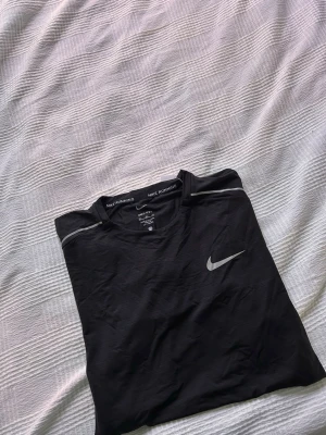 Svart Nike Running t-shirt - Svart t-shirt från Nike Running med rund hals och diskreta grå detaljer på axlarna. Tillverkad i lätt och snabbtorkande Dri-FIT-material som passar perfekt för löpning och träning. Stor Nike-logga på bröstet. Storlek Xxl men sitter som M