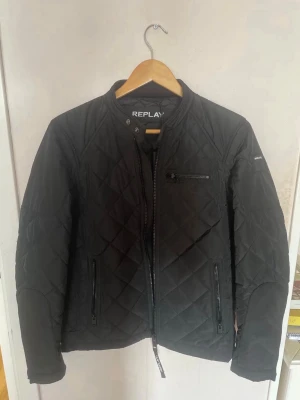 Svart quiltad pufferjacka Replay - Stilren svart pufferjacka från Replay med quiltat mönster och dragkedjor framtill. Jackan har en smal passform, ståkrage och flera praktiska fickor med dragkedja. Perfekt för kyliga dagar när du vill ha en snygg och enkel look.