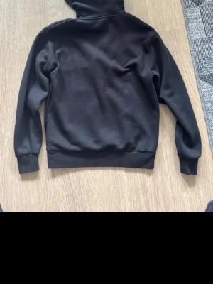 Svart Hoodrich hoodie med tryck - Svart hoodie från Hoodrich med stort grafiskt tryck i vitt och rött på bröstet samt mindre tryck på huvan. Klassisk känguruficka framtill och ribbade muddar vid ärmslut och midja. Perfekt för en streetwear-look.