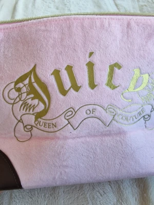 Rosa Juicy Couture make up bag - Söt rosa kuvertväska från Juicy Couture med mjuk sammetstextur och guldigt tryck på framsidan. Väskan har mörkbruna hörn i läderimitation och dragkedja upptill. Perfekt accessoar för att sticka ut med en lyxig och trendig vibe.