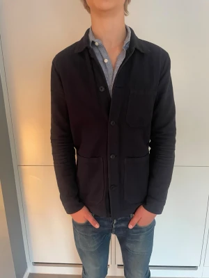  Mörkblå over shirt från A days March - Stilren mörkblå overshirt från A days match med klassisk krage och tre stora fickor framtill. Overshirten har knäppning med knappar och är tillverkad i mjuk bomull. Perfekt att bära över en skjorta för en avslappnad men snygg look.