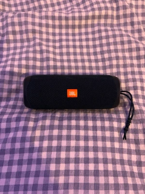 Jbl Flip 5 högtalare - Snygg svart bärbar högtalare från JBL med robust tygmaterial och gummidetaljer. Kompakt och cylinderformad design med orange JBL-logga på framsidan och praktisk snodd för enkel transport. Perfekt för att ta med sig musiken överallt.