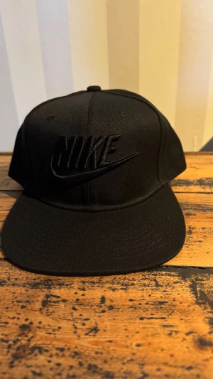 Svart keps från Nike med broderad logga - Snygg svart keps från Nike med broderad logga framtill i samma färg för en clean look. Klassisk modell med böjd skärm och justerbar baksida. Perfekt accessoar för en sportig och stilren outfit.