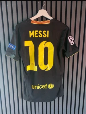 FC Barcelona Messi tröja svart/gul - Säljer en svart FC Barcelona matchtröja med Messi 10 på ryggen. Tröjan har gula detaljer, klubbmärke och Nike-logga framtill samt Champions League-märke på ärmen. Materialet är lätt och andas, perfekt för fotbollsträning eller match.