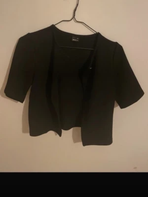 Svart kortärmad kofta från Gina Tricot - Stilren svart kortärmad kofta från Gina Tricot. Koftan har öppen front och rak passform, vilket gör den enkel att matcha med olika outfits. Perfekt för lager på lager och funkar året runt. Materialet känns mjukt och bekvämt.