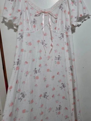 Vitt nattlinne med rosett och mönster - Sött vitt nattlinne med rosa blommor och grå figurer. Lång modell med korta ärmar och en liten rosett framtill. Tillverkat i mjukt och bekvämt material, perfekt för mysiga kvällar hemma.