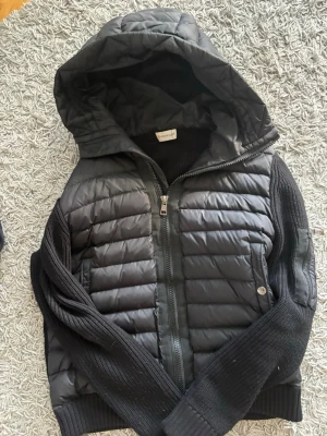 Svart dunväst med huva från Moncler - Snygg svart dunväst från Moncler med stickade svarta ärmar och huva. Västen har dragkedja framtill och Moncler-logga på ena ärmen. Perfekt för lager-på-lager och streetstyle. Materialmix av dun och stickat ger en cool look.