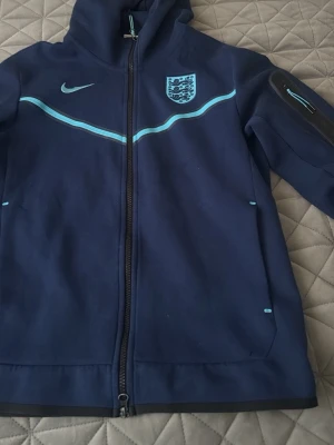 Nike tech england - Snygg mörkblå hoodie från Nike med Englands landslagslogga på bröstet och ljusblå detaljer. Dragkedja framtill, huva och fickor på sidorna. Tillverkad i mjukt material, perfekt för dig som gillar sportig stil. Den är sällsynt och sälls inte mer bra skick  tecken på användning finns men bra skick